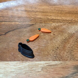 May include: Three cookie cutters on a wooden surface. Two are orange carrot shaped cutters with green tops. One is a black, detailed, carrot shaped cutter.