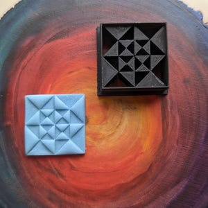 Puede incluir: Dos diseños geométricos impresos en 3D, uno en negro y otro en azul claro, sobre un fondo de madera con un degradado arcoíris.