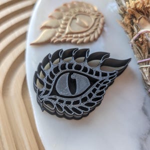 May include: A black plastic dragon eye cookie cutter with intricate details. The cutter is shaped like a dragon eye with a sharp point at the end.