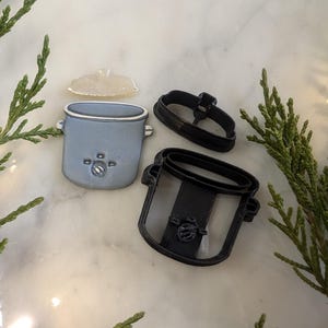 Puede incluir: Un juego de dos cortadores de galletas con forma de olla de cocción lenta con tapa. Los cortadores son negros y de plástico. La olla de cocción lenta es de color azul claro y tiene una tapa blanca.