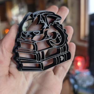 Puede incluir: Cortador de galletas de plástico negro con forma de dragón sobre una pila de libros. El dragón tiene un diseño detallado y los libros tienen lomos visibles. Ideal para hornear y manualidades.