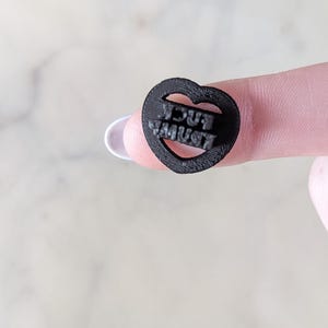 May include: A small, black heart-shaped object with the words "YOU MUST" cut out. The object is held between two fingers against a light background.