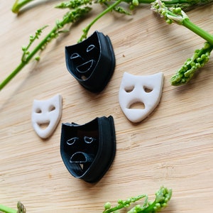 Puede incluir: Cuatro cortadores de galletas con forma de máscaras teatrales: dos negros y dos blancos. Los cortadores negros representan una cara sonriente y una cara triste. Los cortadores blancos muestran una cara sonriente y una cara triste. Ramitas verdes en el fondo.