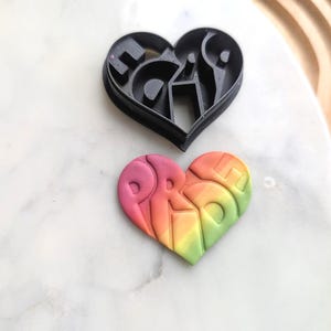 May include: A black heart-shaped cookie cutter with the word "PRIDE" cut out. A rainbow-colored heart-shaped cookie with the word "PRIDE" embossed on it.