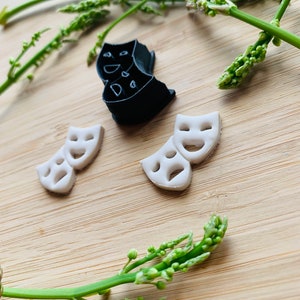 May include: A black plastic theater mask cookie cutter and two small white clay theater mask charms. Each charm features a pair of comedy and tragedy masks with cut-out eyes and mouths. The items are arranged together on a light wood surface.