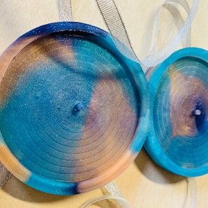 Puede incluir: Dos posavasos redondos de resina de color azul verdoso, marrón y morado con un patrón de remolino. Los posavasos tienen un centro elevado y un acabado liso y brillante.