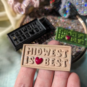 May include: A tan-coloured rectangular sign with the text "MIDWEST IS BEST" in raised letters. A red heart is between the words "IS" and "BEST".