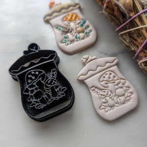 May include: Two ceramic mushroom jar ornaments, one white with painted details and one black with a raised design. A black plastic cookie cutter in the shape of a mushroom jar is also pictured.
