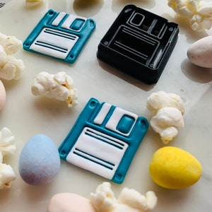May include: Two blue and white floppy disk shaped cookies and a black floppy disk shaped cookie cutter on a white surface with popcorn and candy eggs.
