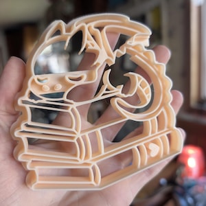 Puede incluir: Un cortador de galletas de plástico beige con forma de dragón posado sobre una pila de libros. El dragón tiene detalles intrincados, incluyendo una cola curvada y alas. El cortador se sostiene en una mano.