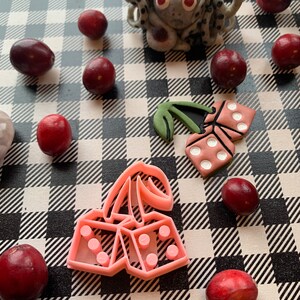 Puede incluir: Cortadores de galletas de plástico rosa en forma de cerezas con dados en la parte superior, y un solo dado con un tallo de cereza en la parte superior. Los cortadores están sobre una superficie a cuadros en blanco y negro con arándanos rojos.