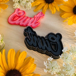May include: Two cookie cutters, one pink and one black, both shaped like the word "Girl" in a cursive font. The cutters are on a wooden surface with yellow flowers around them.