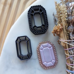 May include: Three decorative gem-shaped cookie cutters. Two are black and one is a light pink color with a gold border.