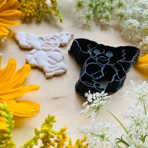 Peut inclure: Deux emportes-pièces en forme de koala. L'un est blanc et l'autre est noir. Les emportes-pièces sont sur une surface en bois entourée de fleurs jaunes et blanches.