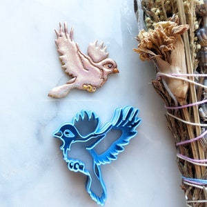 May include: Two bird cookie cutters, one is light blue and the other is light pink with gold accents. The cookie cutters are on a white marble surface.