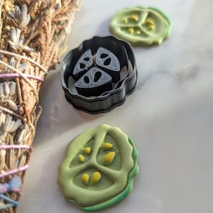 Puede incluir: Un cortador de galletas de plástico negro con forma de rodaja de pepino con tres semillas. También se muestran dos rodajas de pepino de fondant verde y amarillo.
