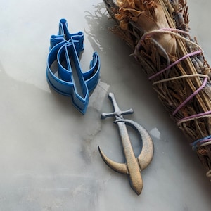 Puede incluir: Dos cortadores de galletas en forma de media luna con una espada en su interior. Un cortador es de plástico azul y el otro es dorado.