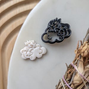 Puede incluir: Dos cortadores de galletas en forma de nube, uno negro y otro blanco, sobre una superficie blanca. Los cortadores están hechos de plástico y tienen un diseño detallado.