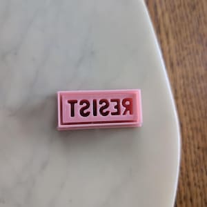 May include: A pink rectangular cookie cutter with the word "RESIST" in a reverse font. The cutter is placed on a white and grey marble surface.