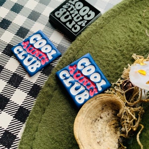 May include: Two blue and white square pins with the text "Cool Aunts Club" in a retro font. The pins are on a green and white checkered surface.