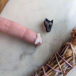 May include: Two small bee-shaped cookie cutters, one black and one white, on a marble surface. A bundle of dried herbs is in the background.