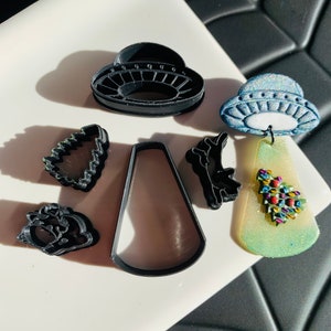 May include: A set of seven black plastic cookie cutters, including a UFO, a Christmas tree, a star, a bell, and a cone. The cutters are on a white surface.