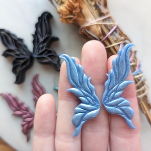 May include: A pair of blue fairy wings made of polymer clay. The wings have a textured, iridescent finish.