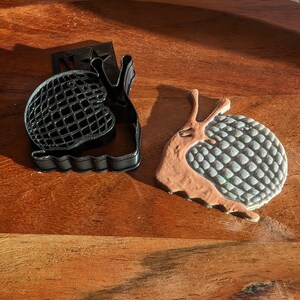 May include: Two snail-shaped cookie cutters, one black and one brown with a blue and white shell. The black cutter has a star on the handle.
