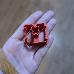 May include: A red plastic cookie cutter in the shape of the letter "M" with a decorative floral design in the center. The cutter is held in a person's hand, with a wooden surface in the background.