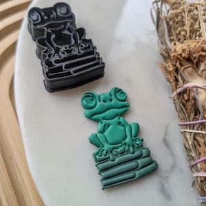 May include: Two frog cookie cutters, one black and one green, both sitting on a stack of books. The cutters are made of plastic and are designed to cut out cookies in the shape of a frog sitting on a stack of books.