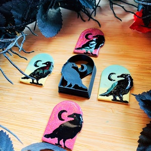 May include: Five black crow silhouette ornaments with a crescent moon on a wooden background. The ornaments are in different colors: pink, blue, and green. One ornament is a 3D printed crow silhouette.