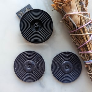 May include: Three black circular coasters with a concentric circle design. The coasters have a speckled black finish.