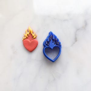 Puede incluir: Un cortador de galletas de plástico azul en forma de corazón con llamas en la parte superior. Junto a él hay una galleta roja en forma de corazón con llamas amarillas.