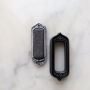 May include: Two black metal frames with ornate designs. The frame on the left has a black background with silver glitter. The frame on the right is empty.