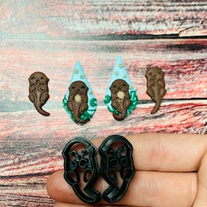 Puede incluir: Una colección de suministros de manualidades con temática de nutria. Incluye dos recortes marrones con forma de nutria, dos adornos azules en forma de lágrima con diseños de nutria y dos cortadores de galletas negros con forma de nutria. Sobre una superficie de madera.