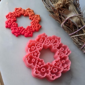 May include: Two pink and orange floral wreaths made of polymer clay. The wreaths are on a white marble surface.