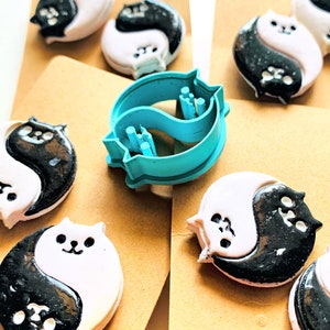 May include: A set of six black and white yin yang cat cookie cutters, with one blue yin yang cat cookie cutter. The cookie cutters are on a brown surface.