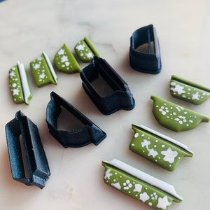 Puede incluir: Un conjunto de nueve cortadores de galletas, incluyendo cuatro cortadores con patrones verdes y blancos y cinco cortadores negros. Los cortadores tienen forma de cuencos y rectángulos.