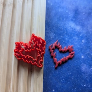 May include: Two red plastic cookie cutters shaped like hearts with a flame design. The cutters are on a wooden surface and a blue background.