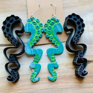 May include: A pair of green and blue octopus-shaped earrings with silver hooks. The earrings are made of polymer clay and have a marbled effect.