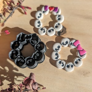 May include: Three cookie cutters in the shape of a circle of skulls with a bow on top. The cutters are made of black plastic and are on a wooden surface.