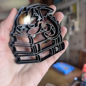 Puede incluir: Cortador de galletas de metal negro con forma de dragón posado sobre una pila de libros. El dragón tiene alas detalladas y una cola curvada. Los libros tienen lomos y páginas visibles. El cortador se sostiene en una mano.