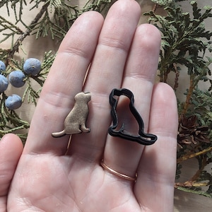 May include: Two dog-shaped cookie cutters. One is gold-colored, the other is black. The gold-colored dog is solid, while the black dog is an outline. The cutters are small and likely used for baking. The background includes greenery.