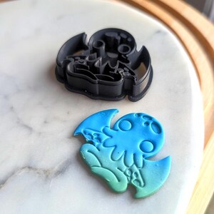 May include: Two cookie cutters in the shape of a cartoon octopus. One is black and the other is blue and green.