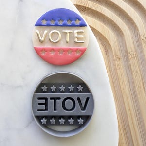 Peut inclure: Deux emportes-pièces, l'un rouge, blanc et bleu avec le mot "VOTE" en lettres dorées, et l'autre gris avec le mot "VOTE" en lettres inversées.