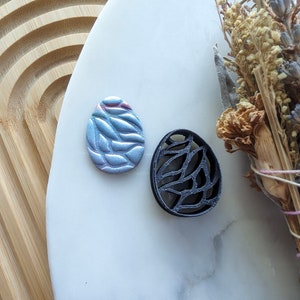 May include: Two decorative round ornaments with a leaf pattern. One ornament is silver with a blue iridescent finish and the other is black.