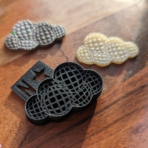 May include: Three cloud-shaped objects with a grid pattern. One is a black plastic frame with a star cutout, one is a shimmering silver piece, and one is a translucent cream-colored piece. Each cloud features a textured, rounded surface design.