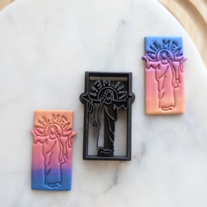 May include: Three cookie cutters, one black and two pink and blue, with the image of a person with a halo and the words "All Men" around the image.