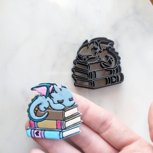 Puede incluir: Una figurita de dragón azul y gris durmiendo sobre una pila de libros coloridos. El dragón tiene alas rosas y una cola amarilla. Los libros están apilados uno encima del otro y tienen diferentes colores y patrones.