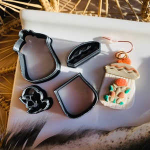May include: A set of five black plastic cookie cutters shaped like a mug, a burger, a bottle, a ghost, and a small round shape. The cutters are on a white surface. There is also a small, handmade clay ornament with a brown, white, and orange design.